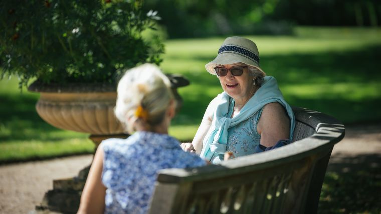 Two elderly women, one with her back to us, one facing us, sitting on a curved stone bench in the Oxford Botanical Gardens