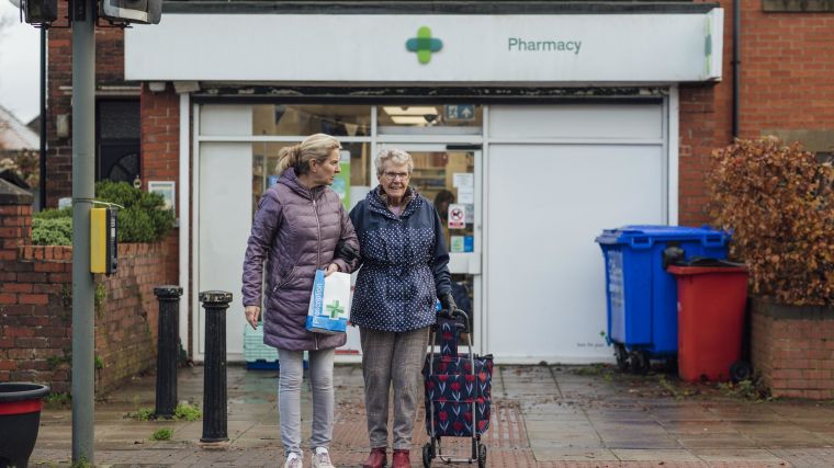 Image of two women leaving a pharmacy shop with medication in a bag - an older woman with a shopping bag on wheels is helped by a younger woman,