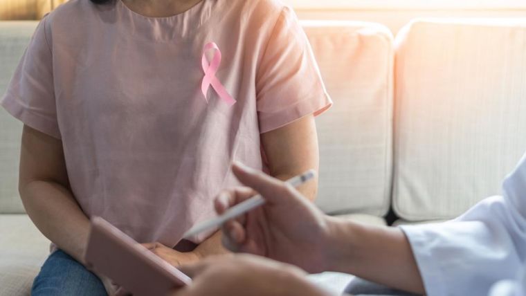 Photo of a woman with a breast cancer awareness pink ribbon sat on a sofa and in consultation with a doctor who is making notes.