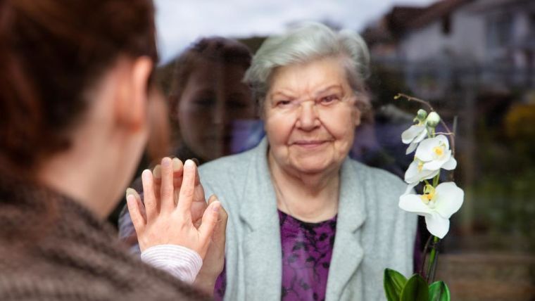 An older lady indoors with her hand up to the glass. A younger person makes a heart sign at her.