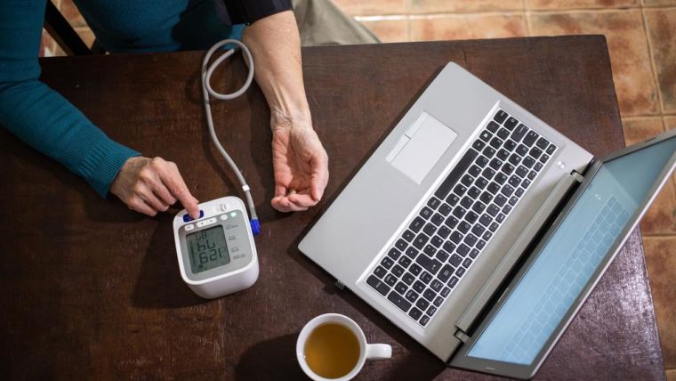 A lady measuring her blood pressure at home using a cuff, and a laptop.